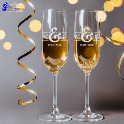 Dois copos de champagne gravados com & M&G 11/06/2022 com fitas douradas ao redor num fundo cinzento com luzes amarelas.