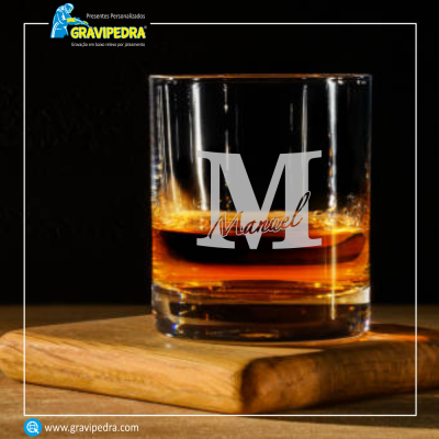 Copo de vidro transparente com bebida âmbar e gravação 'M Manuel' sobre tábua de madeira