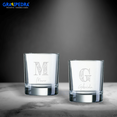 Dois copos de vidro personalizados com letras e nomes, sobre superfície cinzenta