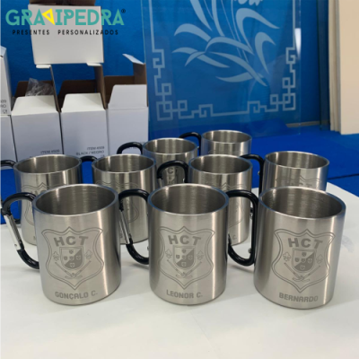 Conjunto de canecas metálicas prateadas gravadas com brasão e nomes, sobre mesa branca em ambiente azul.