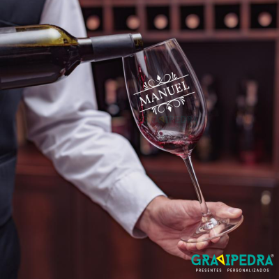 Taça de vinho com o nome MANUEL sendo servida num ambiente com garrafas ao fundo