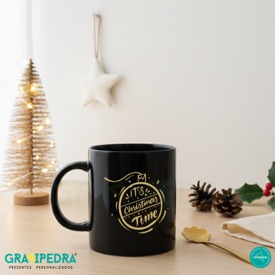 Caneca preta com texto dourado de Natal numa mesa de madeira clara com decoração natalícia