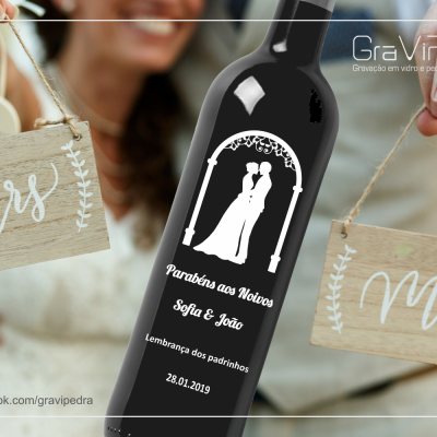 Garrafa preta de vinho comemorativa de casamento com duas placas de madeira 'Mr' e 'Mrs' seguradas por mãos.