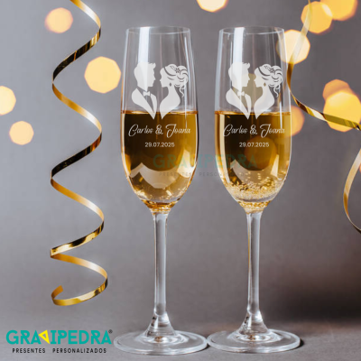 Dois copos de champanhe gravados com silhuetas de casal e datas, decoração festiva com fitas douradas.