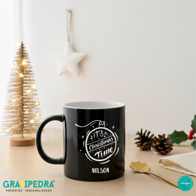 Caneca preta de cerâmica com decoração natalícia e texto em branco numa mesa de madeira clara com decoração de Natal