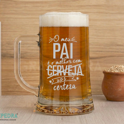 Caneca de vidro com texto branco preenchida com cerveja e espuma, sobre mesa com pote de barro com grãos.