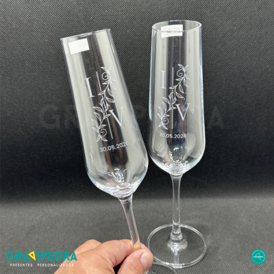 Dois copos de champanhe transparentes com decoração branca e data, fundo preto, mão segurando um copo.