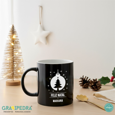 Caneca preta com desenho natalício branco e texto 'FELIZ NATAL MARIANA' sobre mesa com decoração de Natal