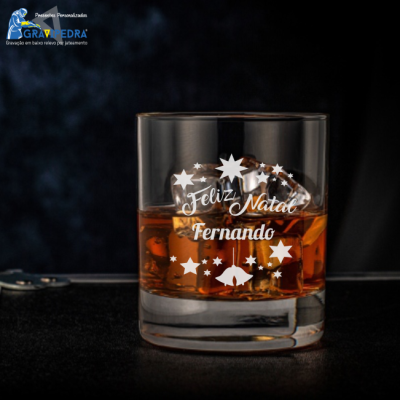 Copo de vidro transparente com whisky, gelo e gravação de Natal personalizada.