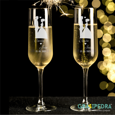 Dois copos de champanhe personalizados com nomes, data e desenho de casal, com faíscas em fundo escuro