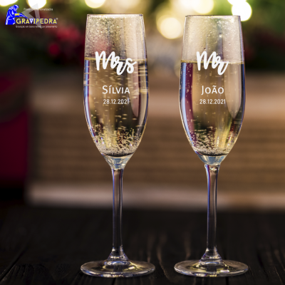 Dois copos de champanhe personalizados com nomes e data, sobre superfície escura e fundo luminoso desfocado.