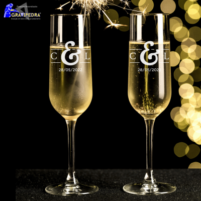 Dois copos de champanhe gravados com letras e data sobre fundo escuro com luzes desfocadas e fogo de artifício