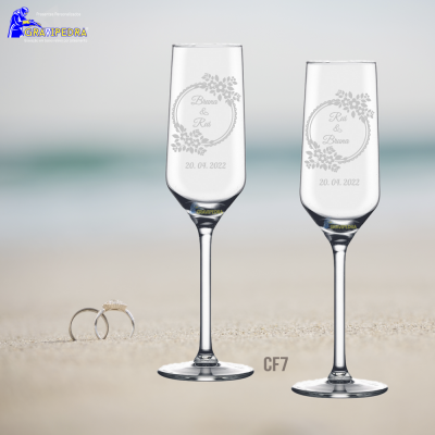 Dois copos de champagne gravados com texto e data, com alianças de casamento na areia e mar desfocado ao fundo.