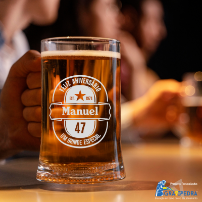 Copo de cerveja personalizado com texto branco em vidro transparente, com cerveja e mão segurando, fundo desfocado