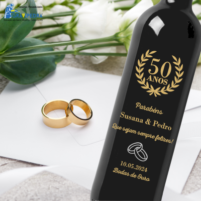 Garrafa preta gravada a dourado para bodas de ouro e dois anéis dourados sobre papel branco e flores.
