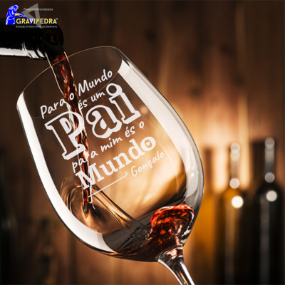 Taça de vinho personalizada com gravação branca, preenchida com vinho tinto, ambiente com garrafas e fundo de madeira.