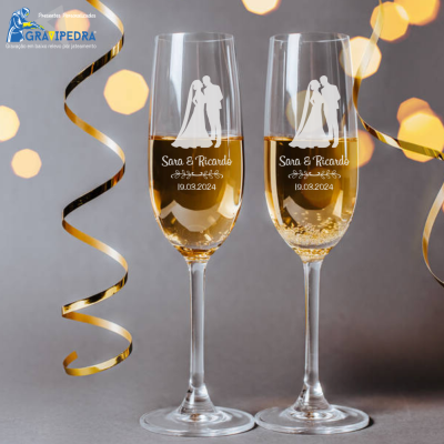 Duas taças de champanhe gravadas com nomes e data num cenário com fita dourada e luzes desfocadas