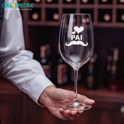 Copo de vidro para vinho com gravação 'PAI', mão segurando, fundo com garrafas