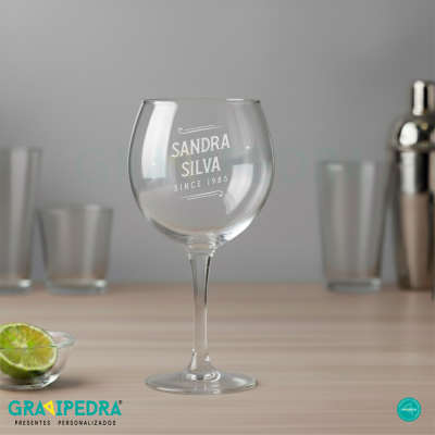 Copo de vidro personalizado com nome e data, sobre mesa de madeira com lima e utensílios para bar ao fundo