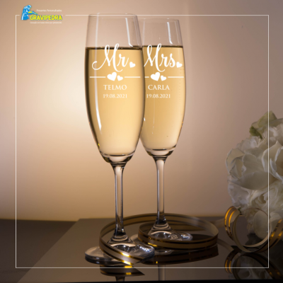 Duas taças de champanhe gravadas com nomes e data aniversários, sobre superfície espelhada, com flores ao lado
