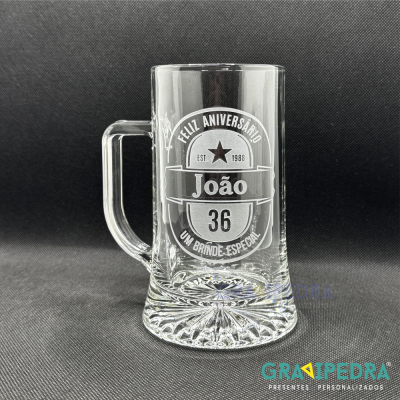 Caneca de vidro com inscrição personalizada para aniversário, sobre fundo preto.