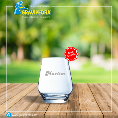 Copo de vidro com nome gravado sobre mesa de madeira e fundo verde desfocado