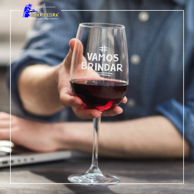 Taça de vinho tinto com texto '# VAMOS BRINDAR' segurada à frente de pessoa com camisa azul