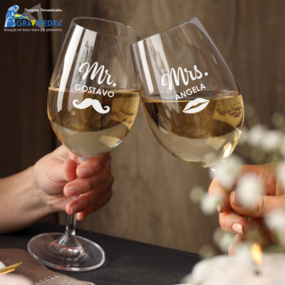 Dois copos de vinho personalizados com nomes e símbolos masculinos e femininos, cheios de vinho branco, sendo erguidos em brindes.