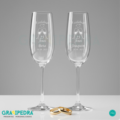 Dois copos de champanhe personalizados com gravações comemorativas e dois anéis dourados numa superfície branca.