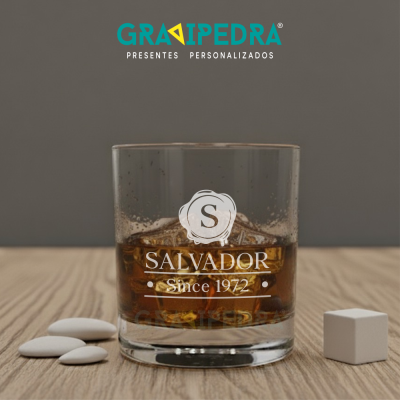 Copo de vidro com bebida castanha, gelo e gravação personalizada, sobre superfície de madeira com pedras e cubo brancos