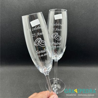 Dois copos de champanhe de vidro transparente gravados com texto e ilustração de alianças, fundo preto.