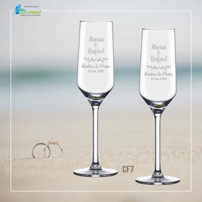 Dois copos de champanhe gravados com nomes e data, anel de noivado ao lado, fundo desfocado