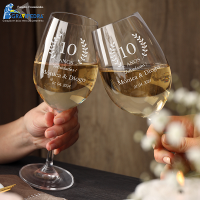 Dois copos de vinho branco gravados a branco com nomes e data, segurados por mãos à mesa
