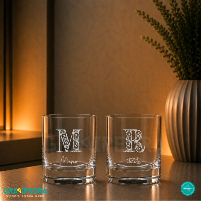 Dois copos de vidro com letras decorativas e nomes Marco e Rute sobre superfície refletora com fundo iluminado e vaso com planta