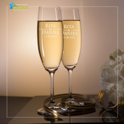 Dois copos de champanhe gravados com nomes e data sobre superfície espelhada