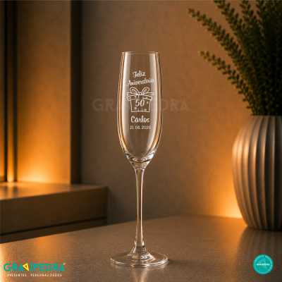 Taça de champanhe gravada com mensagem de aniversário sobre mesa em ambiente iluminado.