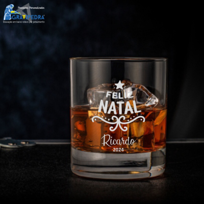 Copo de vidro com bebida âmbar e gelo, com gravação de Feliz Natal 2024