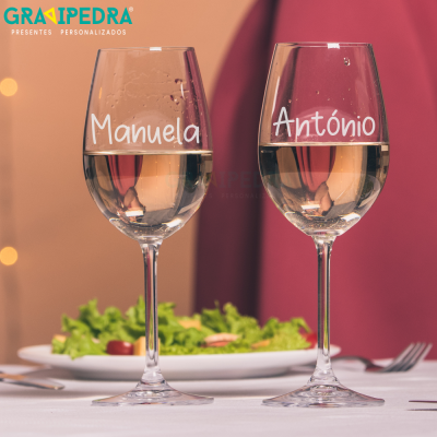 Dois copos de vinho gravados com nomes Manuela e António sobre mesa com comida ao fundo