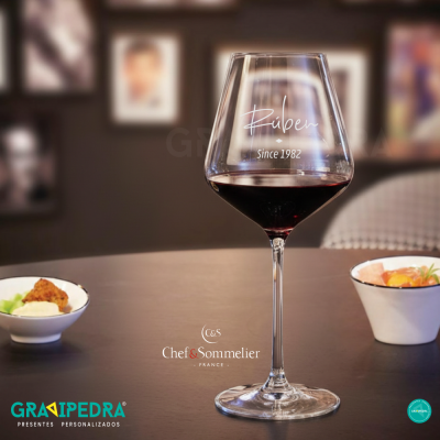 Copo de vinho tinto personalizado com nome Rúben e ano 1982 em mesa com petiscos e quadros ao fundo.