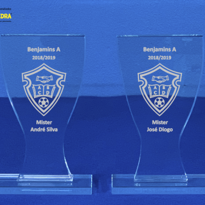 Dois troféus de vidro transparente gravados com textos e brasões sobre superfície azul