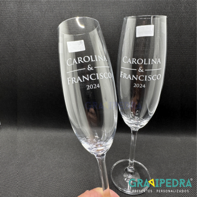 Dois copos de vidro para champanhe com texto branco gravado e etiquetas brancas sobre fundo preto