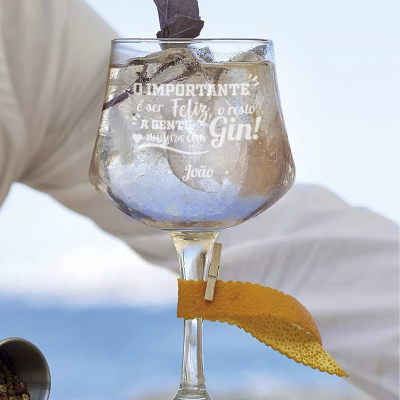 Copo de gin com gelo, folhas e casca de laranja com texto gravado, cenário de mar ao fundo
