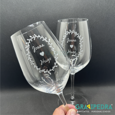 Dois copos de vinho transparentes com nomes gravados Joana e Hugo e um coração entre eles.