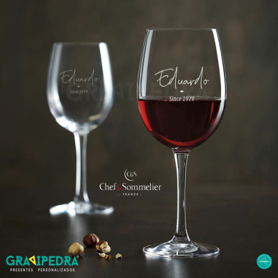 Dois copos de vidro para vinho com gravação personalizada, um cheio de vinho tinto e outro vazio, sobre mesa escura com avelãs à volta