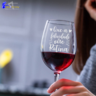 Copo de vinho com gravação branca 'Que a felicidade vire Rotina' e mão a segurá-lo