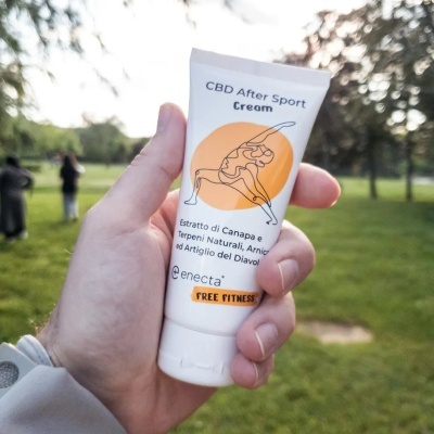 Tubo branco de creme CBD After Sport Cream com design laranja segurado por uma mão em parque ao ar livre
