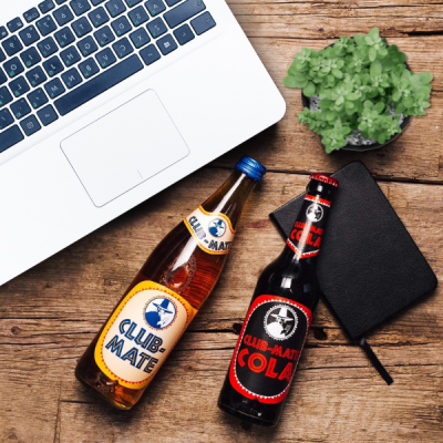 Computador portátil prateado junto a garrafas CLUB-MATE, carteira preta e planta em vaso cinza sobre mesa de madeira