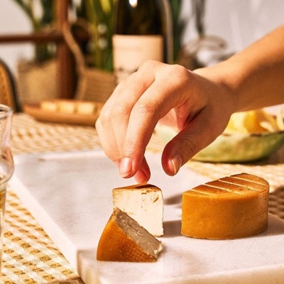 Três pedaços de queijo firme em suporte branco e mão a pegar um pedaço