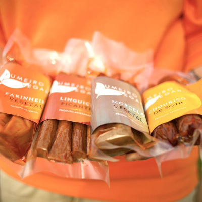 Quatro embalagens de enchidos vegetais coloridos seguradas por pessoa com camisola laranja