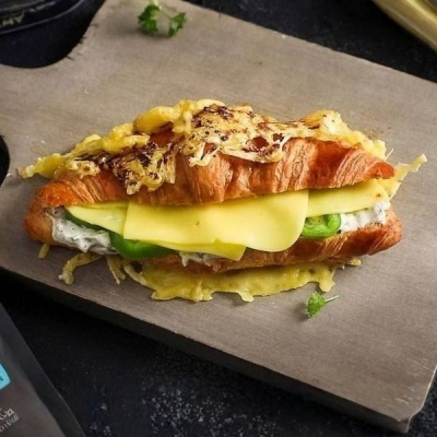 Croissant recheado com queijo, pepino e molho, coberto por queijo gratinado numa tábua